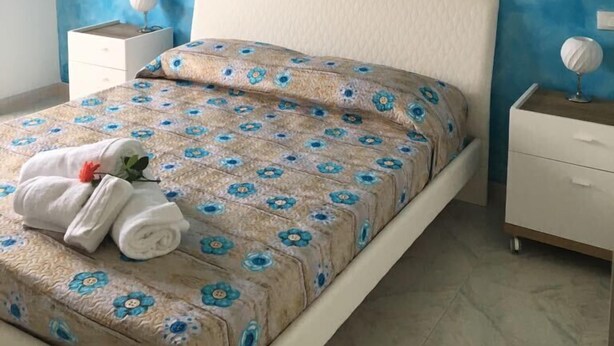 A bedroom with a blue textured wall, a white bed with a floral bedspread, a bedside table with a lamp, and a small globe on the nightstand. - B&B Arcobaleno (Gallipoli)