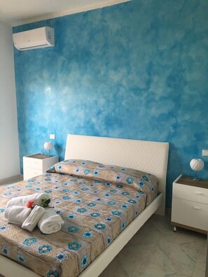 Un dormitorio con pared azul con textura, una cama blanca con colcha floral, una mesita de noche con lámpara y un pequeño globo terráqueo en la mesita.