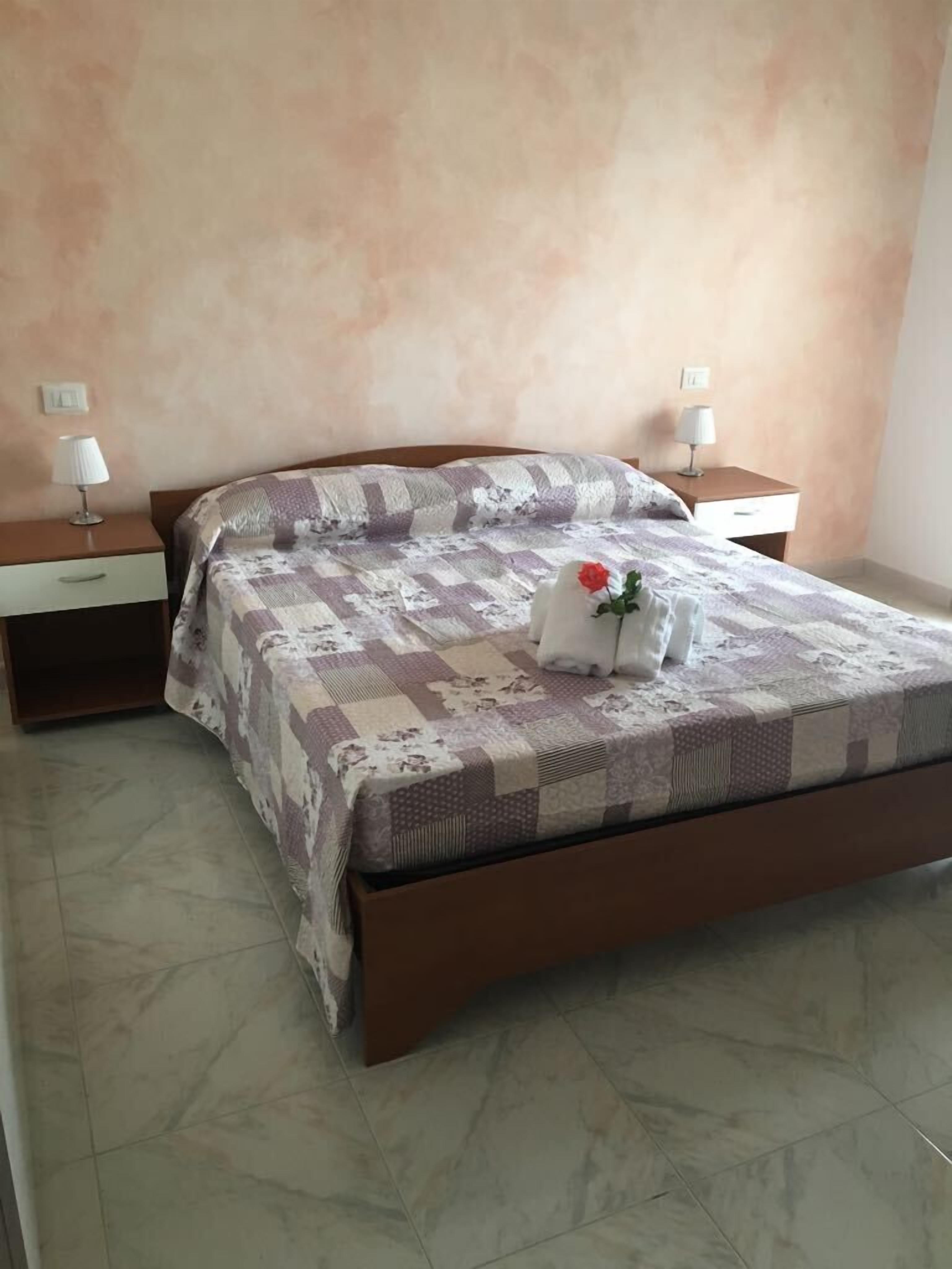 A neatly made bed with a floral-patterned bedspread, two bedside tables with lamps, and a single decorative rose on the bed.