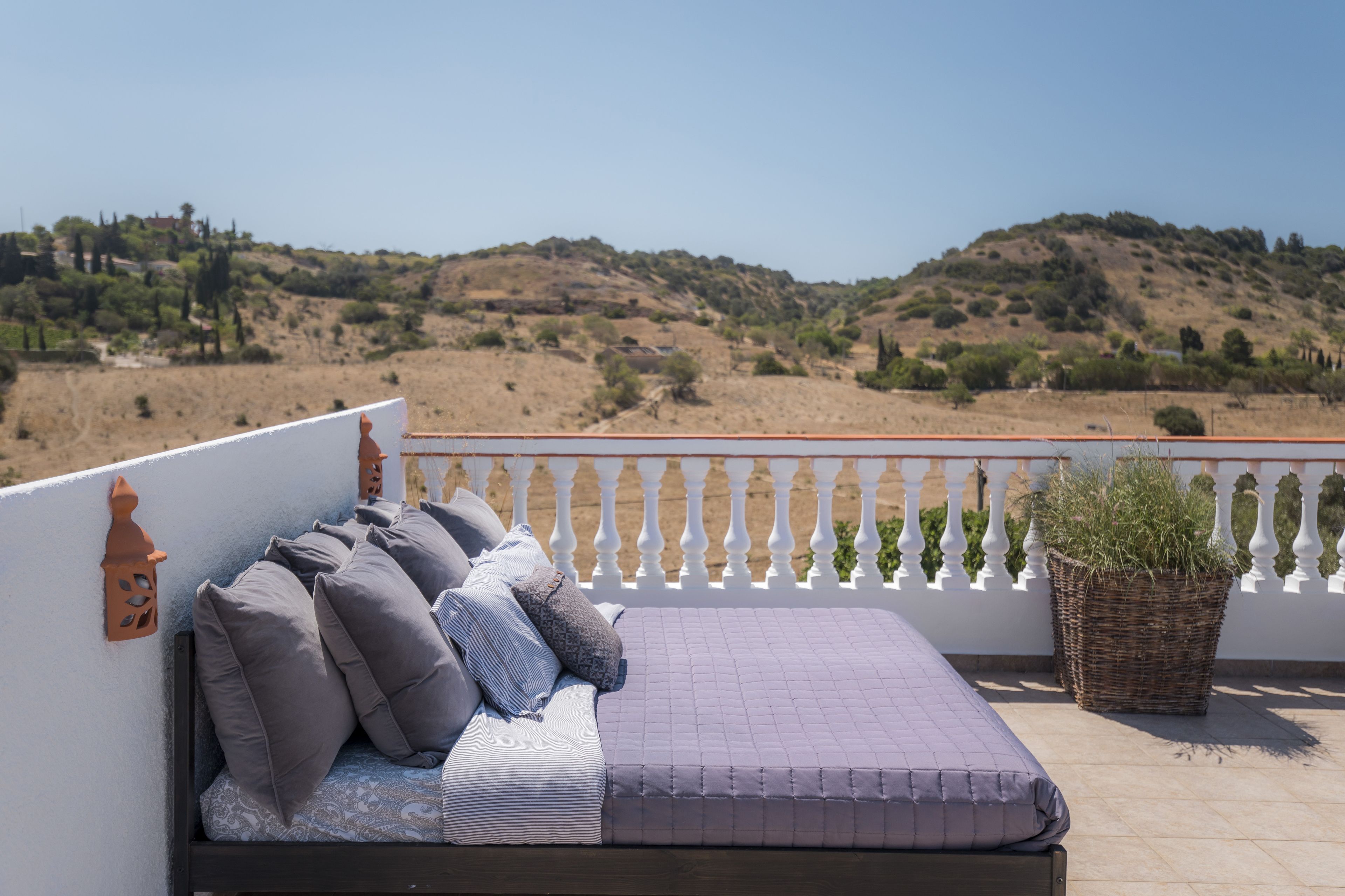 A balcony with a bed, white balustrade, and a view of a hilly landscape.
