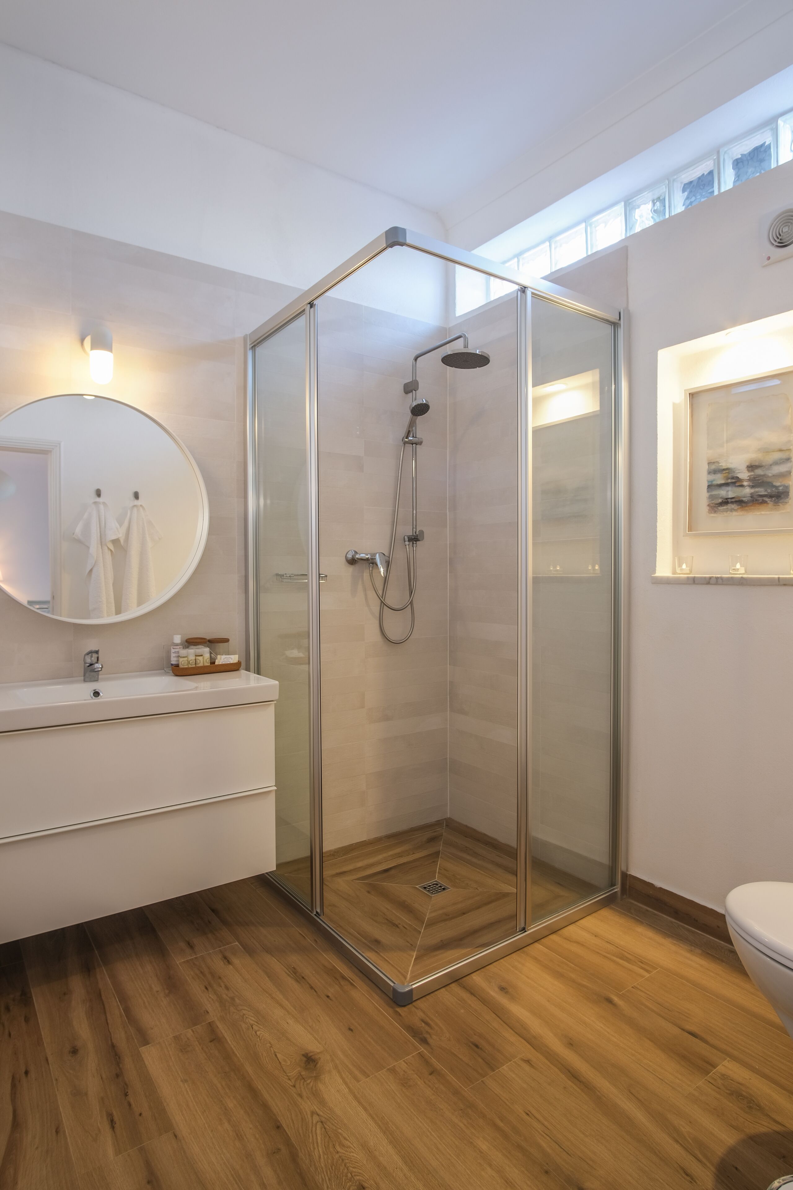 A modern bathroom with a glass-enclosed shower, a round mirror, a white sink, and a wooden floor.
