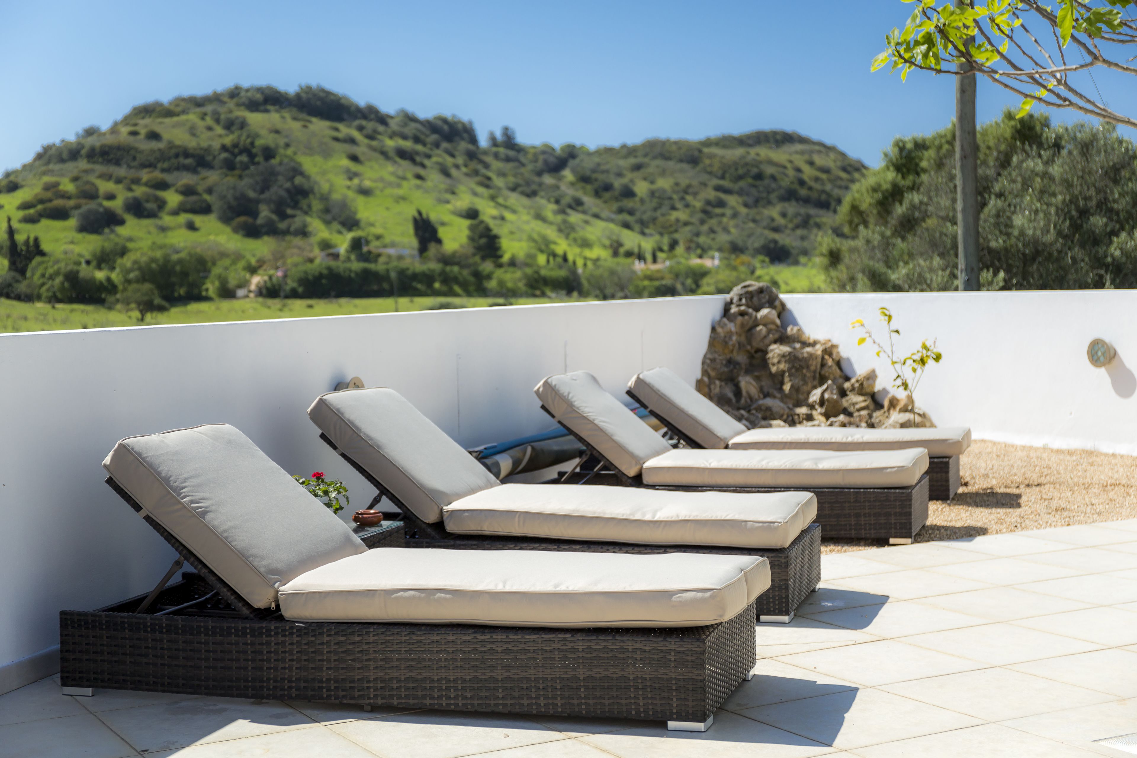 Outdoor wicker lounge chairs with cushions, arranged on a tiled patio with a view of a green hillside.