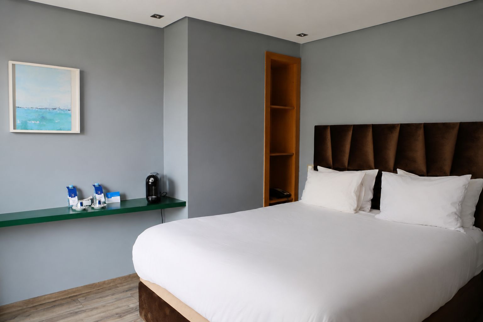 Habitació d'hotel amb un llit, una taula de nit amb una cafetera, una estant amb productes de tocador i un quadre a la paret.