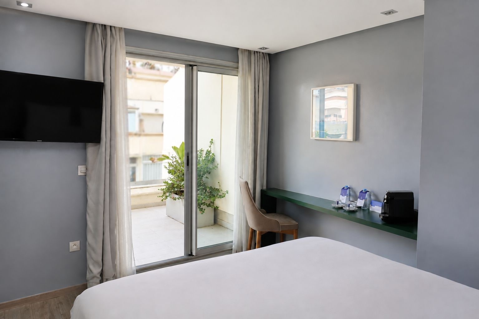 Habitación de hotel con cama, televisor montado en la pared, una mesita con una silla, una planta y un balcón con vistas.