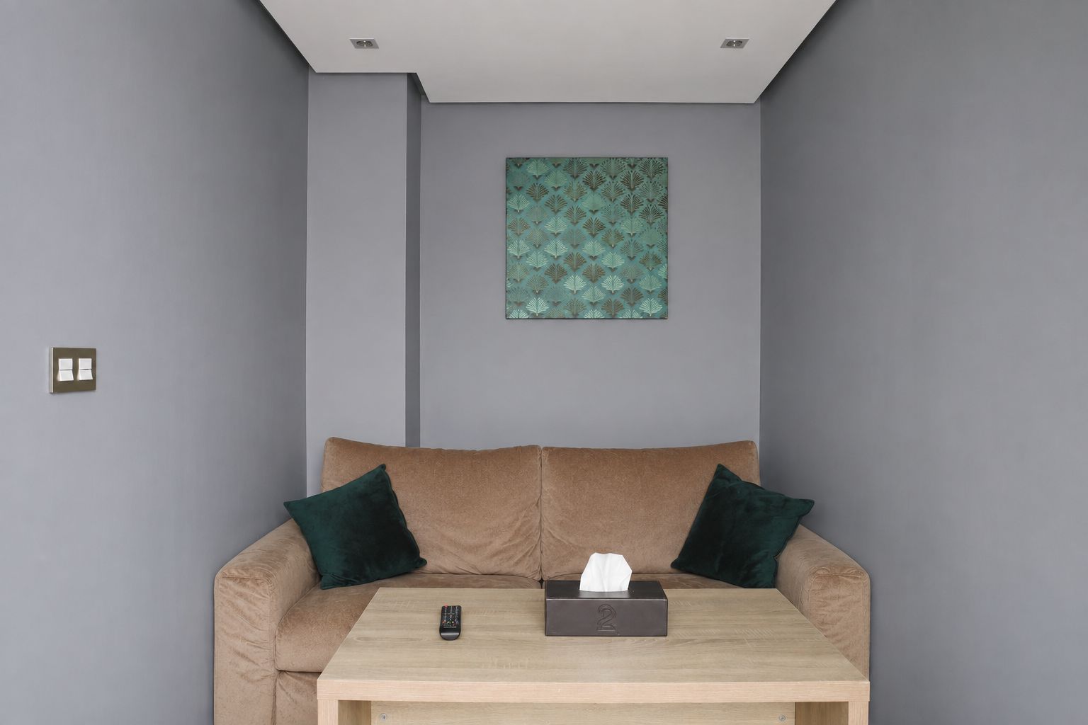 A room with a beige sofa, two dark green pillows, a wooden coffee table, a remote control, and a tissue box.