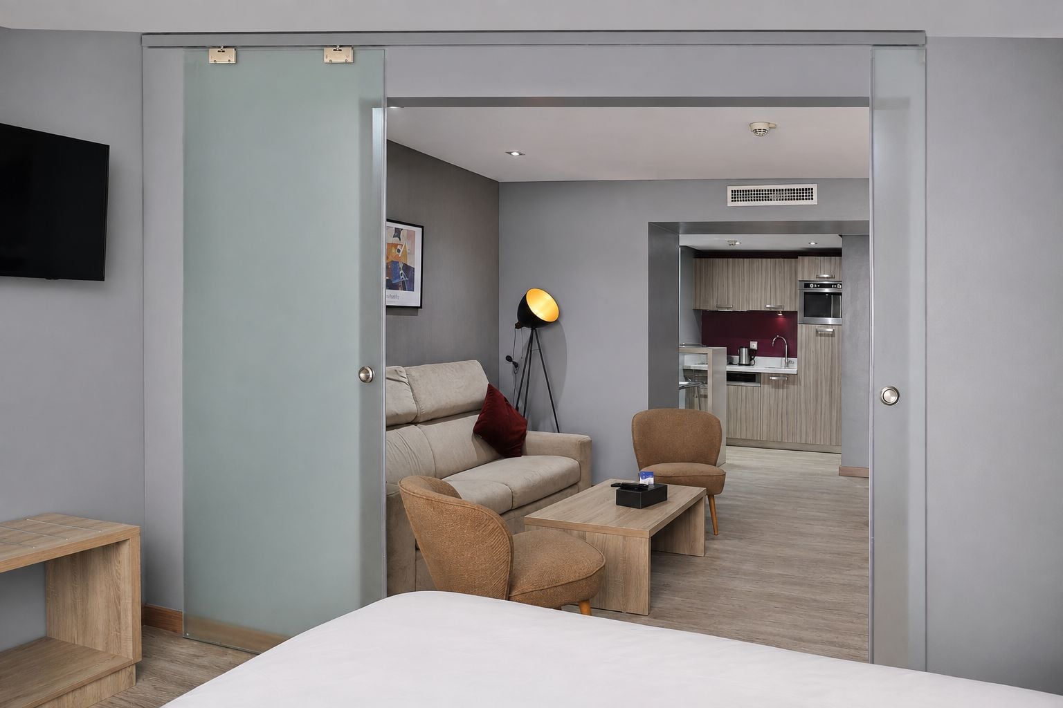 Une chambre d’hôtel moderne, équipée d’un lit, d’un canapé, d’une table basse et d’un bureau. On aperçoit une kitchenette au fond de la pièce.