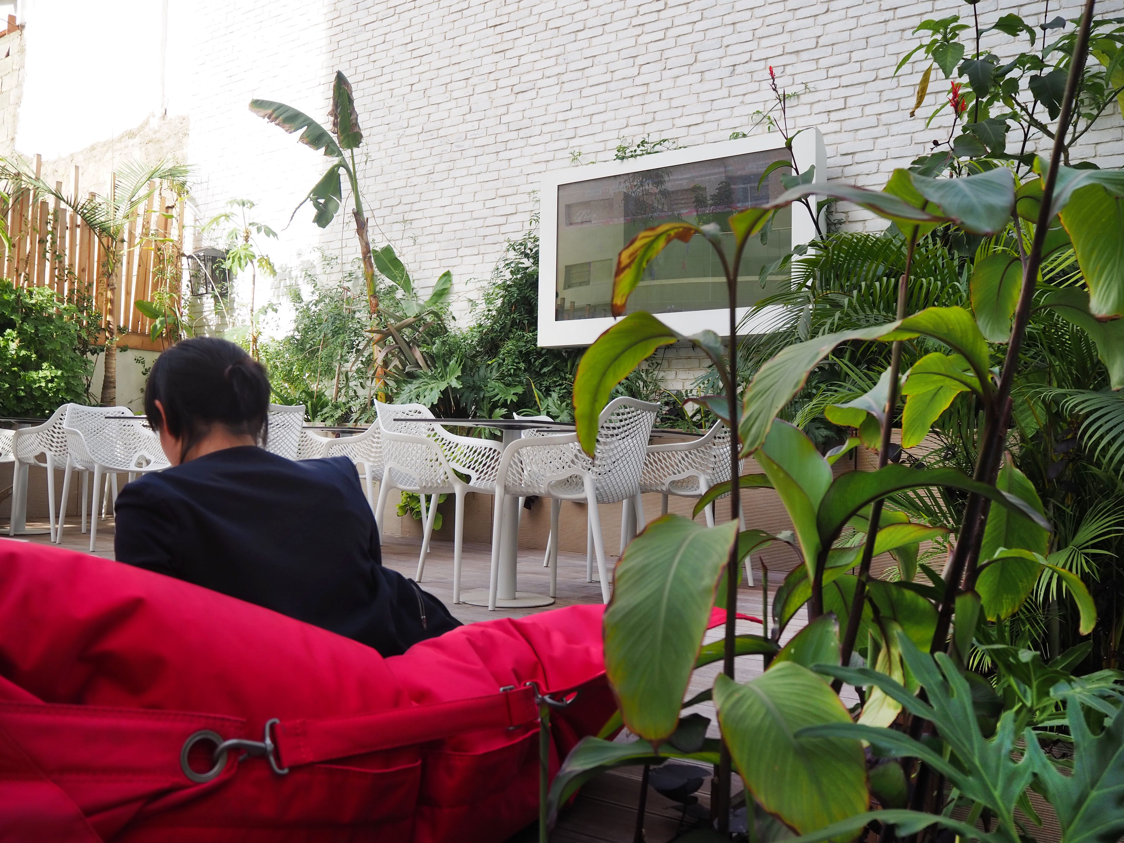 Une personne est assise sur un canapé rouge dans un espace extérieur aménagé en patio, avec des chaises et des tables blanches, entourée d’une végétation luxuriante et d’un mur de briques blanches.