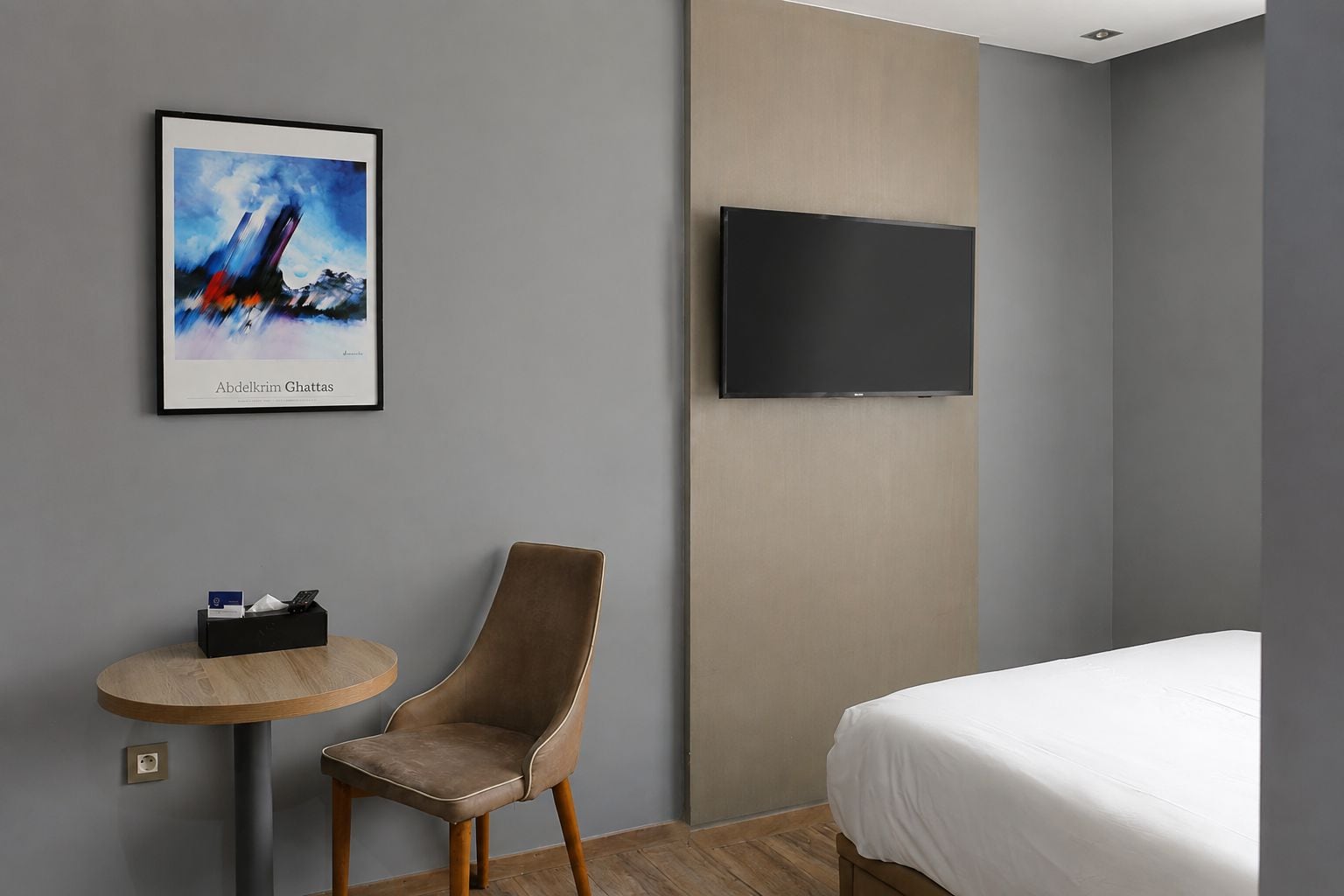 Une chambre d’hôtel avec un lit, une chaise, une petite table, un téléviseur fixé au mur et un tableau encadré accroché au mur.
