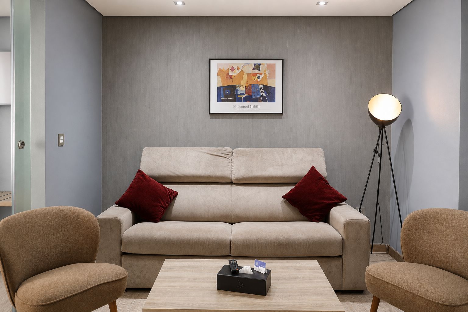Un salon avec un canapé beige, deux coussins rouges, une table basse avec des télécommandes, une œuvre d’art encadrée au mur et une lampe sur pied.