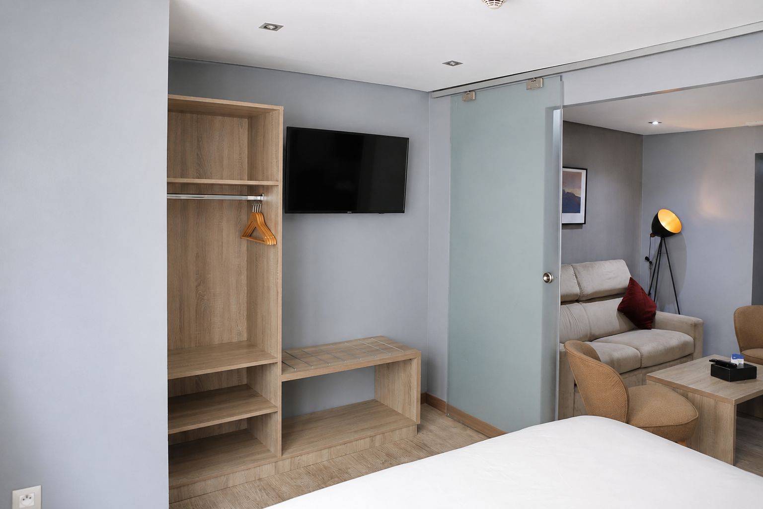 A modern hotel room with a bed, a seating area with a sofa and chairs, a wooden wardrobe, a flat-screen TV, and a floor lamp.