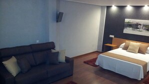 A modern hotel room with a dark-colored sofa, a bed with a white bedspread, a red rug, and a painting on the wall. - Hostal Ciudad de Lepe (Lepe)