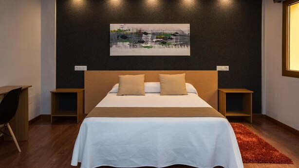 A modern bedroom with a large bed, two bedside tables, a desk, and a painting on the wall. - Hostal Ciudad de Lepe (Lepe)