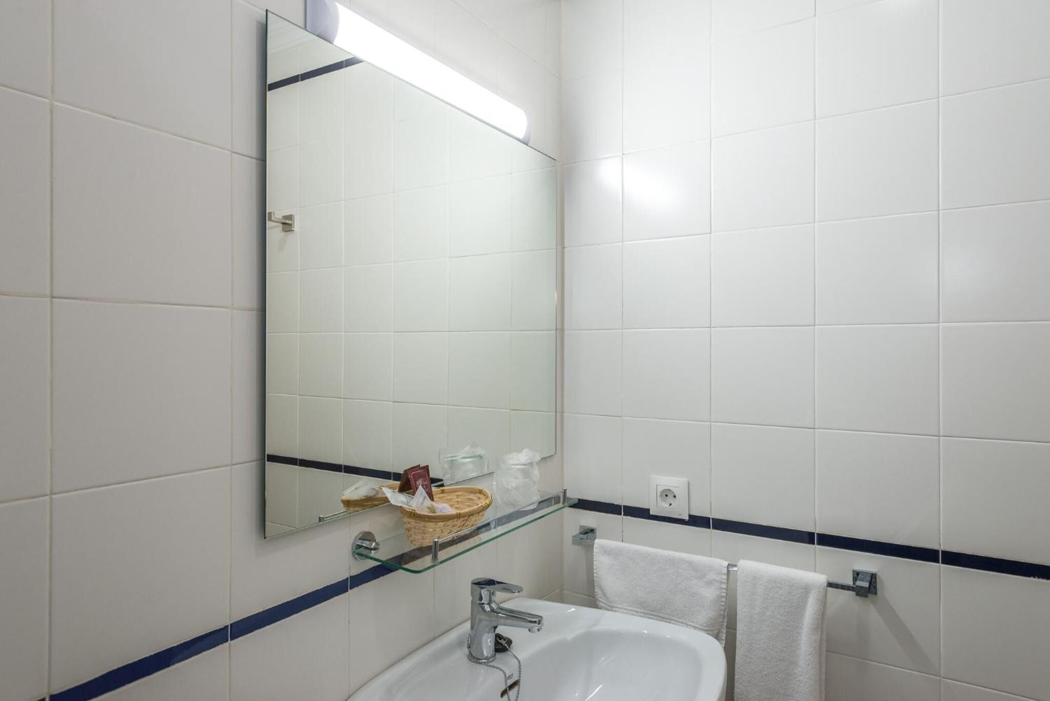 A bathroom with white tiles, a glass shelf with toiletries, a mirror, and a towel rack.