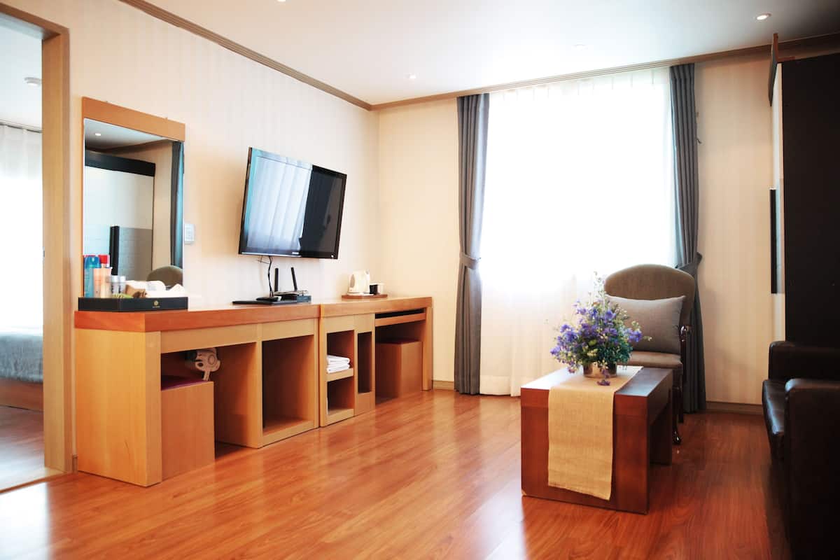 Ein Hotelzimmer mit Schreibtisch aus Holz, einem Flachbildfernseher, einem bequemen Sessel und einem kleinen Tisch mit Blumen.