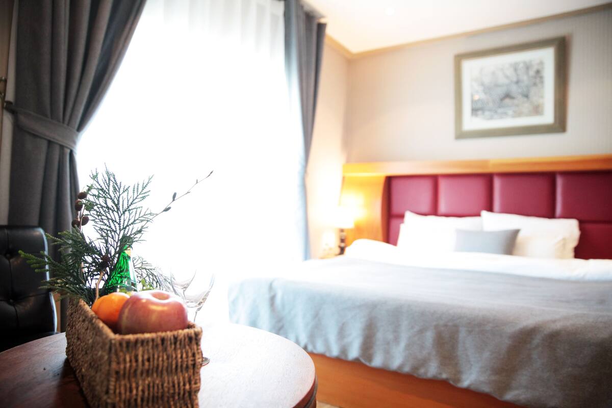 Ein Hotelzimmer mit Bett, Nachttisch, Lampe, einem Korbwagen mit Früchten und einem Fenster mit Vorhängen.