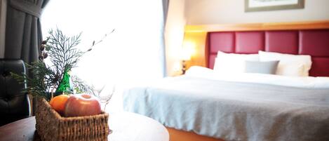 Habitación de hotel con cama, mesita de noche, lámpara, una cesta de mimbre con frutas y una ventana con cortinas.