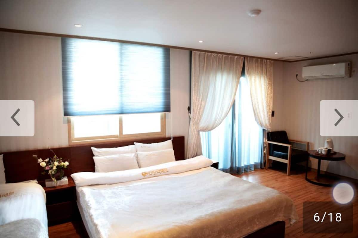 Ein Hotelzimmer mit einem großen Bett, einem Nachttisch mit Blumenvase, einem Schreibtisch mit Stuhl und einem Fenster mit Vorhängen.