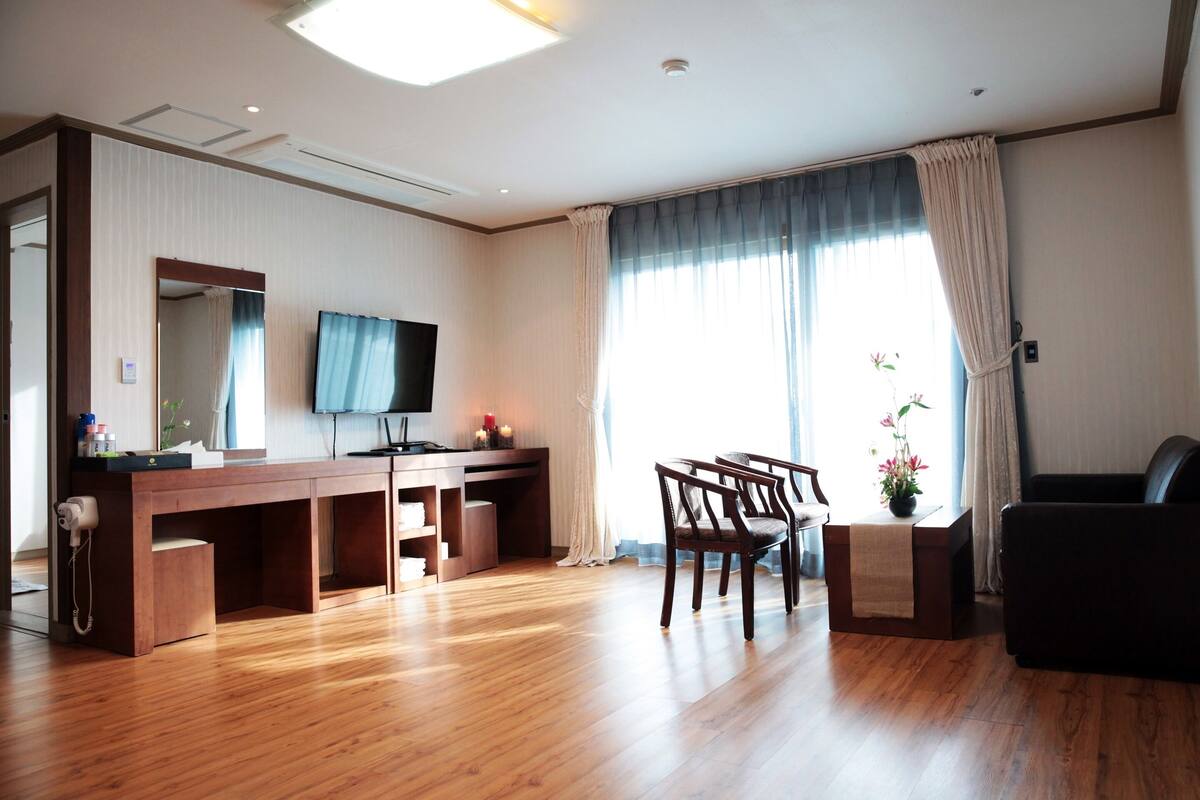 Ein Hotelzimmer mit einem Holztisch, einem Flachbildfernseher, einer Couch und einem großen Fenster mit Vorhängen.