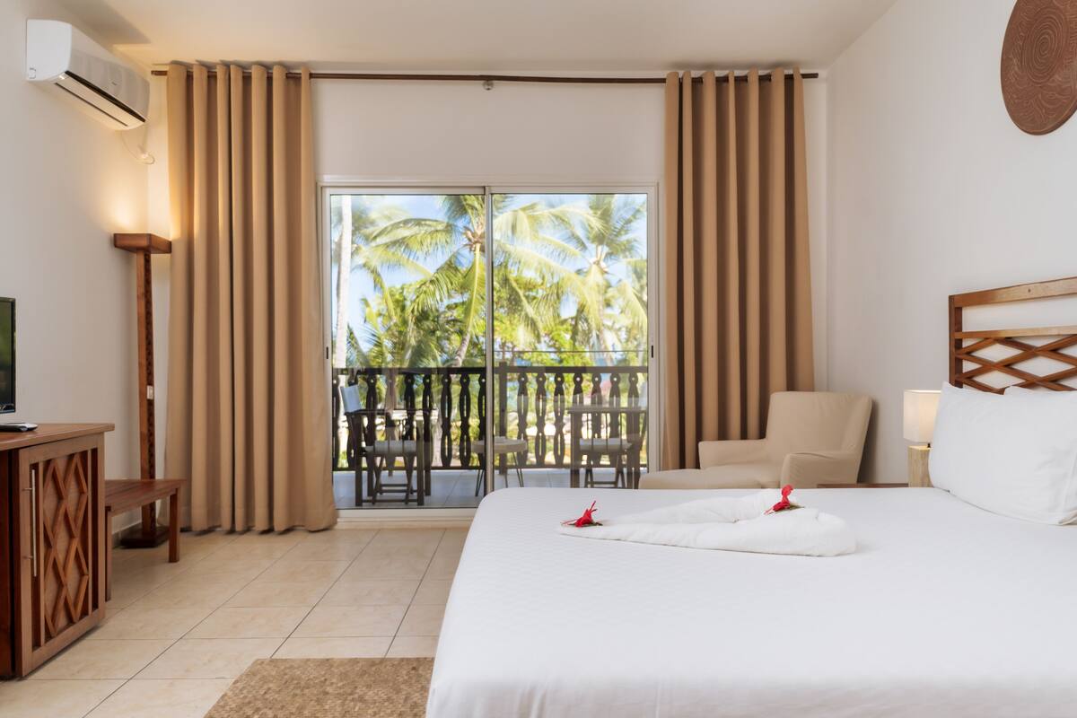 A hotel room with a bed, a chair, a desk, a TV, and a balcony with palm trees.