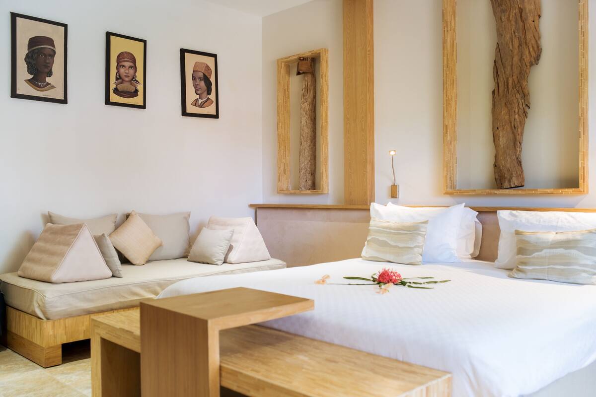 A bedroom with a bed, a sofa, a wooden bench, and three framed artworks on the wall.