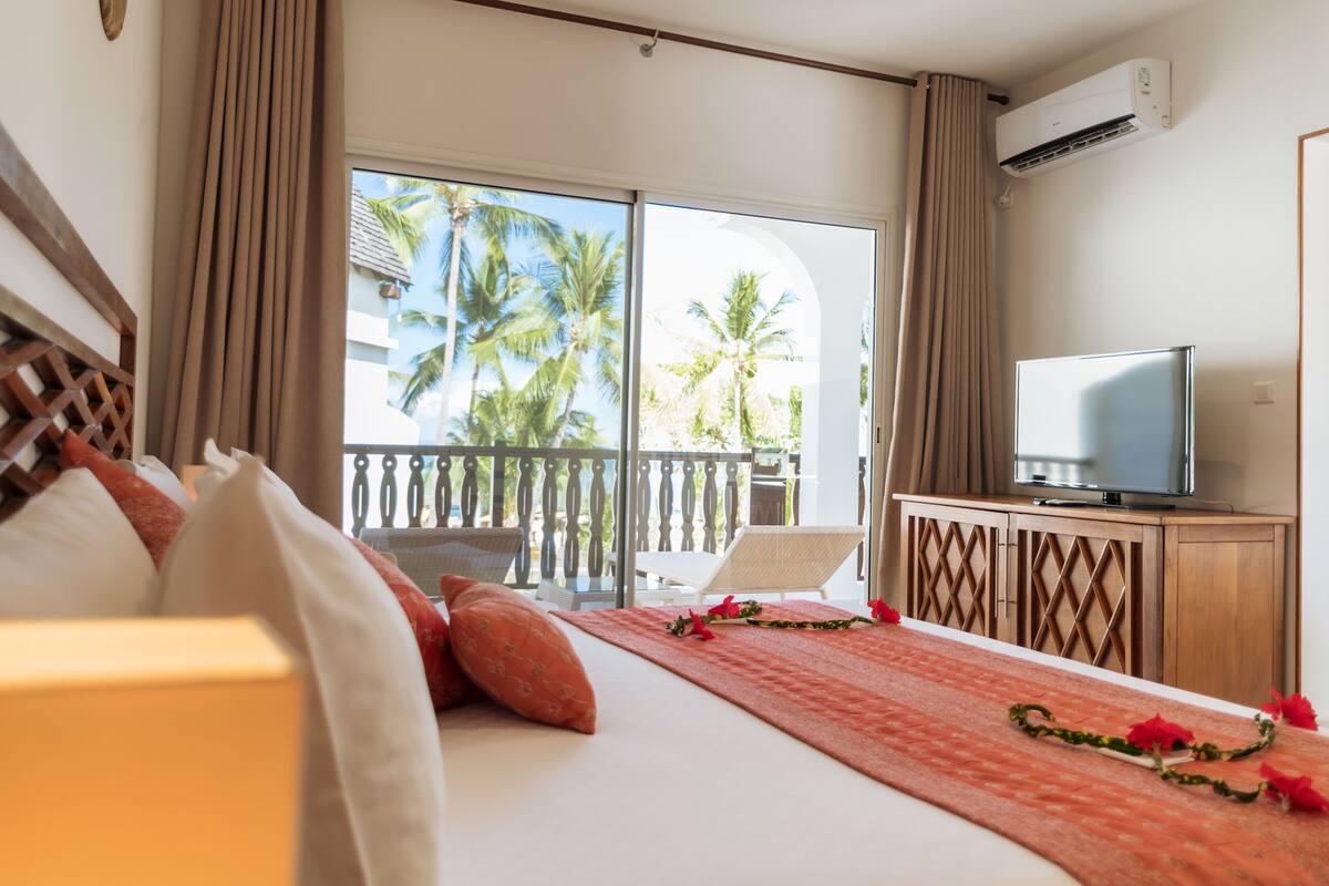 A hotel room with a bed, a TV on a wooden cabinet, a balcony with a view of palm trees, and an air conditioning unit.