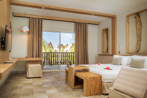 A hotel room with a bed, a desk, a TV, a balcony with a view of palm trees and buildings. - Royal Beach Hôtel (Nosy Be)