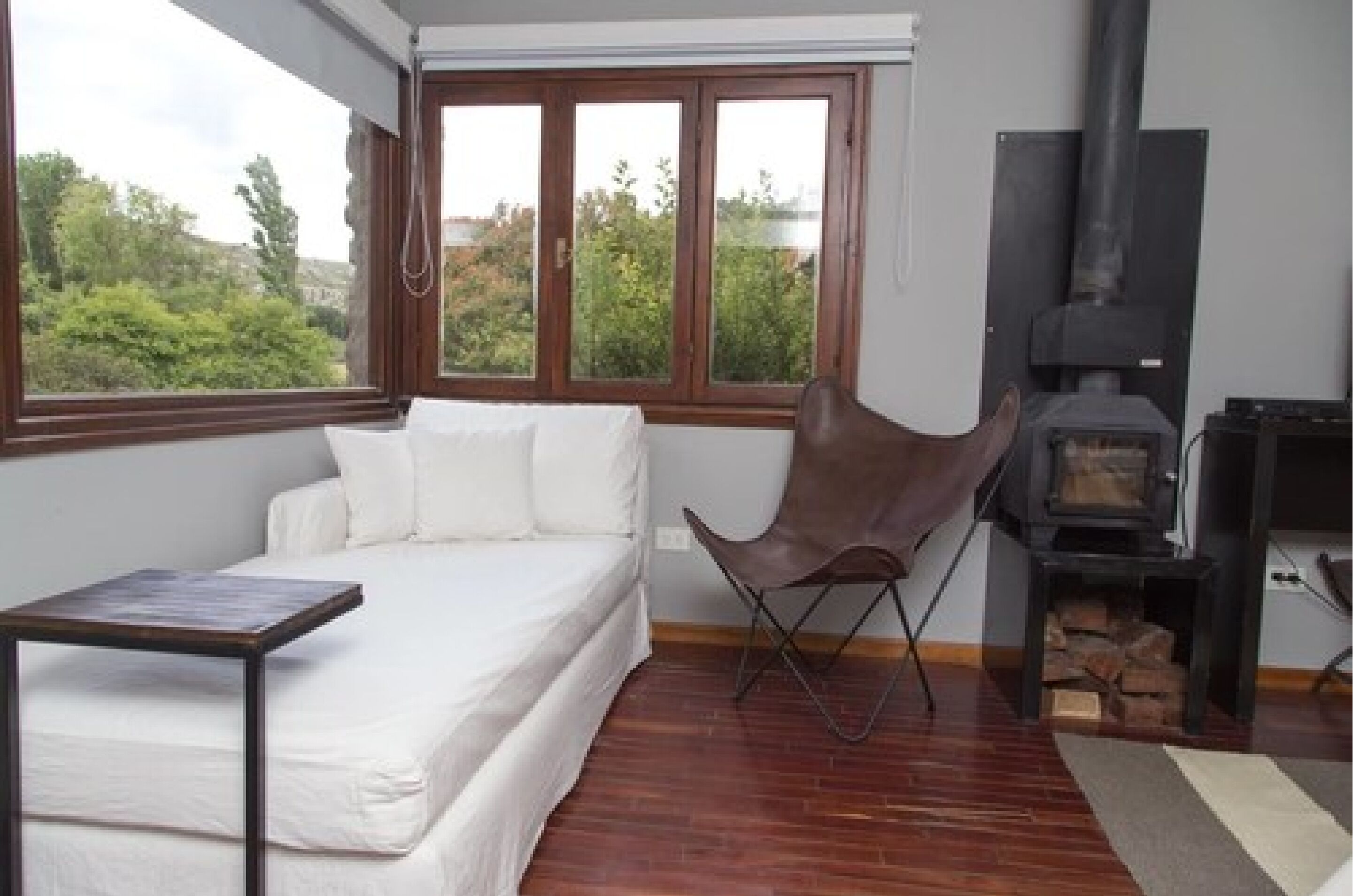 Un dormitorio con cama, una silla, una mesa, chimenea y ventana con vistas a árboles.