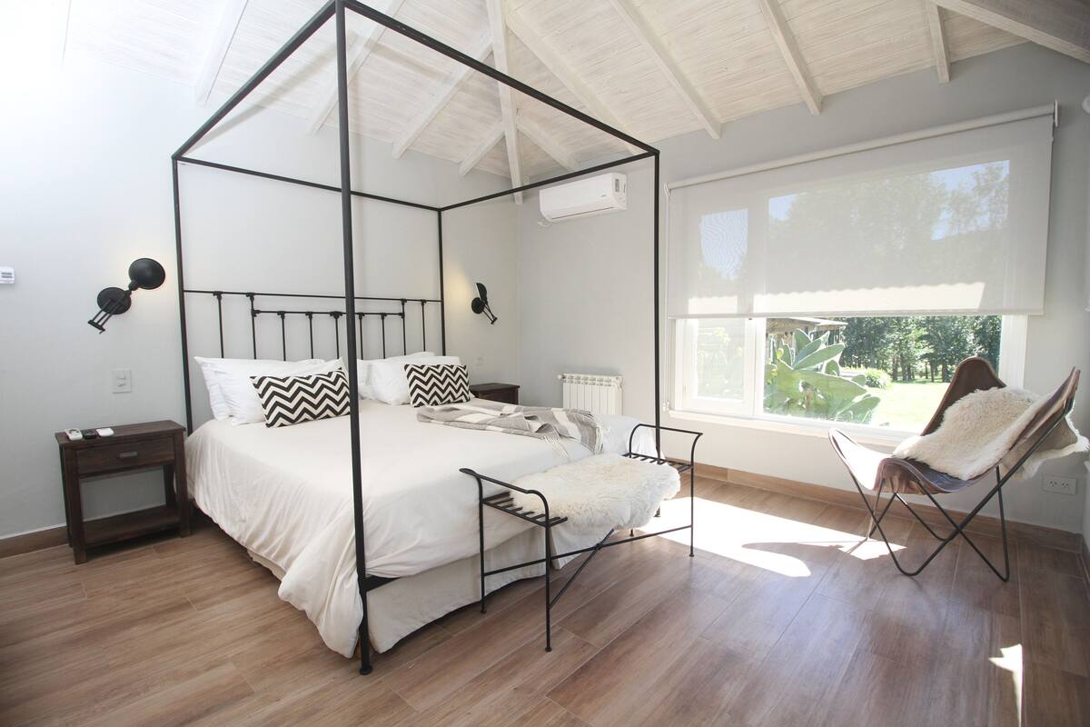 Un lit à baldaquin avec un couvre-lit blanc et des oreillers, une petite table de chevet avec une lampe et une chaise recouverte d’un plaid en fausse fourrure.