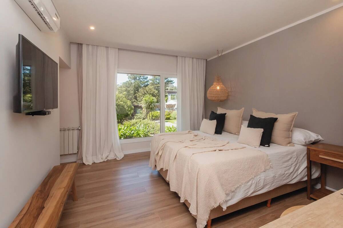 Une chambre avec un grand lit, un téléviseur fixé au mur, une fenêtre avec des rideaux et un plancher en bois.