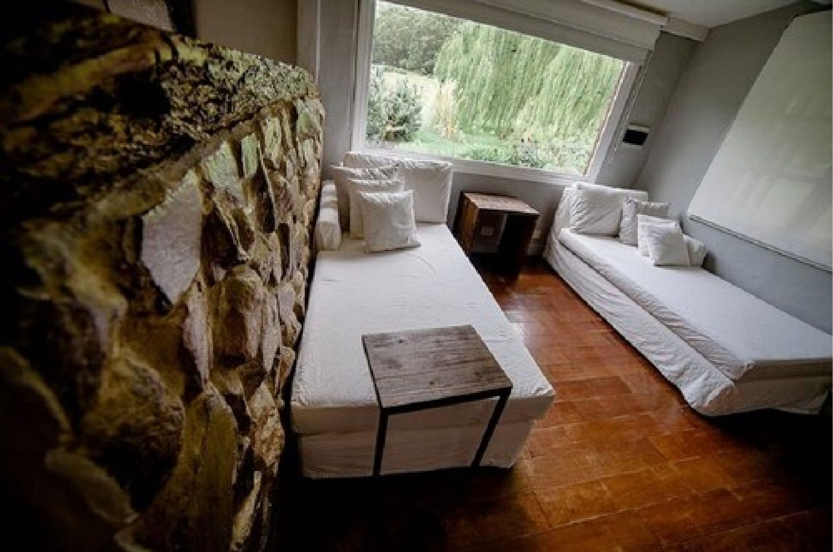 A cozy room with a stone wall, a wooden bench, a cushioned bench, and a window with a view of greenery.
