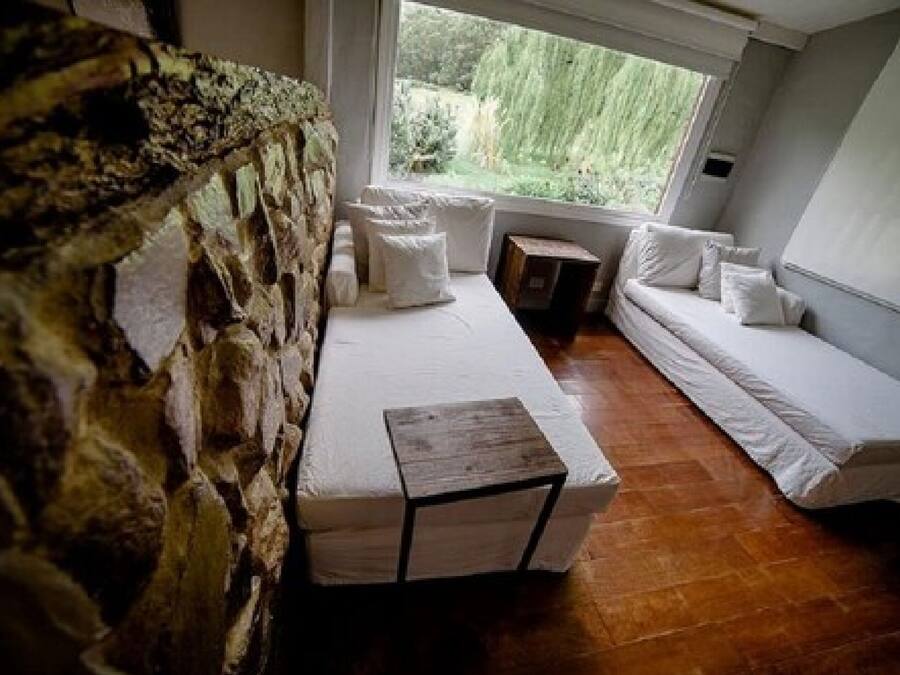 A cozy room with a stone wall, a wooden bench, a cushioned bench, and a window with a view of greenery.