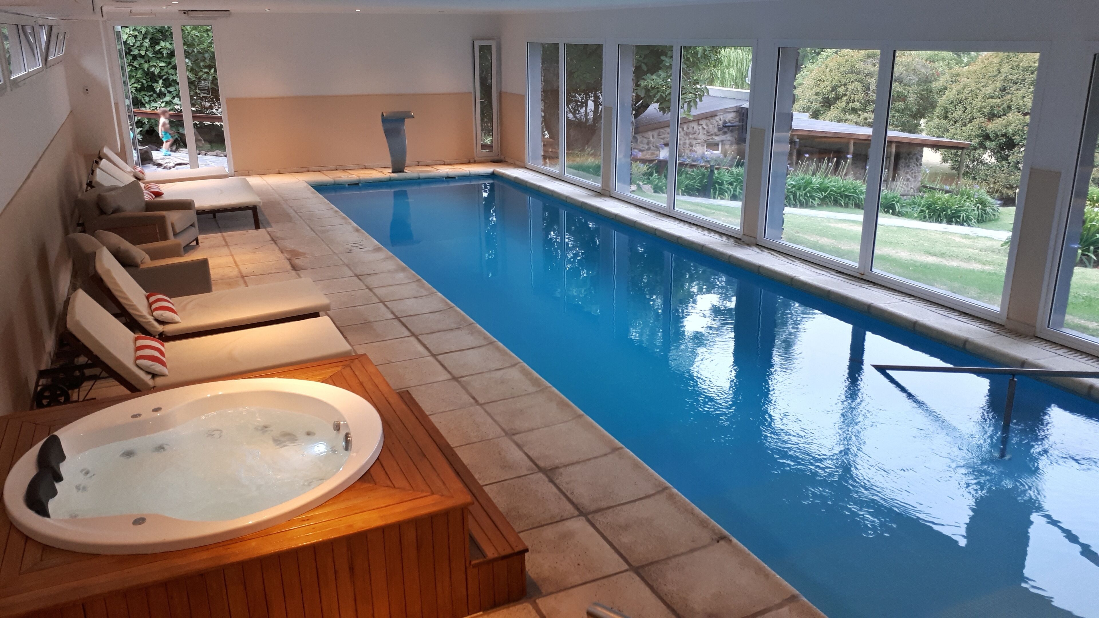 An indoor swimming pool with a jacuzzi, sun loungers, and large windows offering a view of the outdoors.