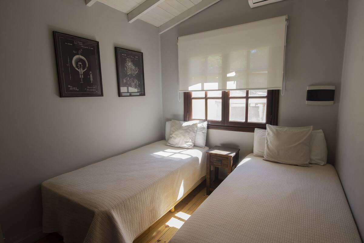 Une chambre avec deux lits simples, une table de chevet, une fenêtre avec des stores et deux certificats encadrés accrochés au mur.