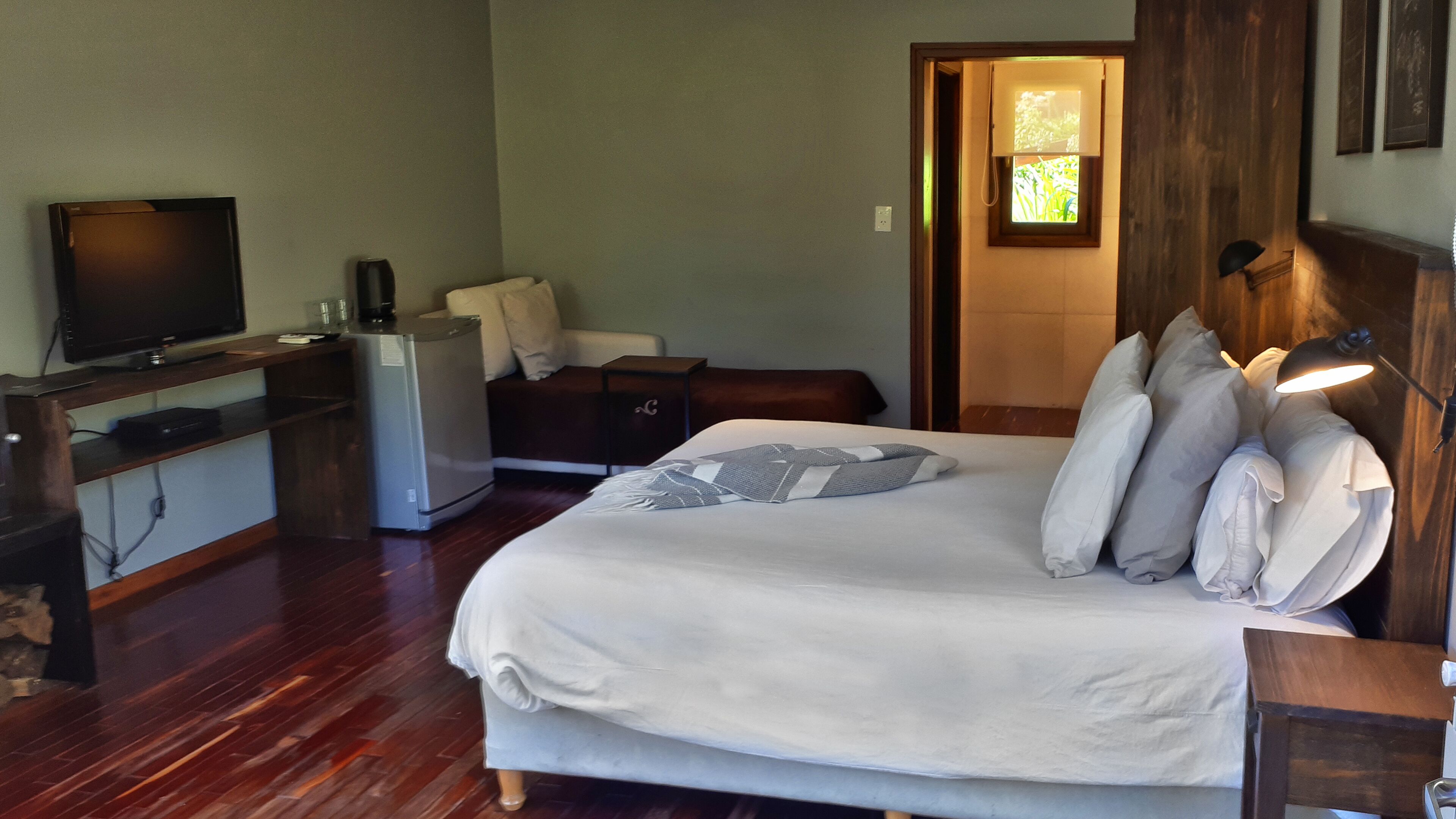 A bedroom with a bed, a desk with a television, a refrigerator, and a window with natural light.