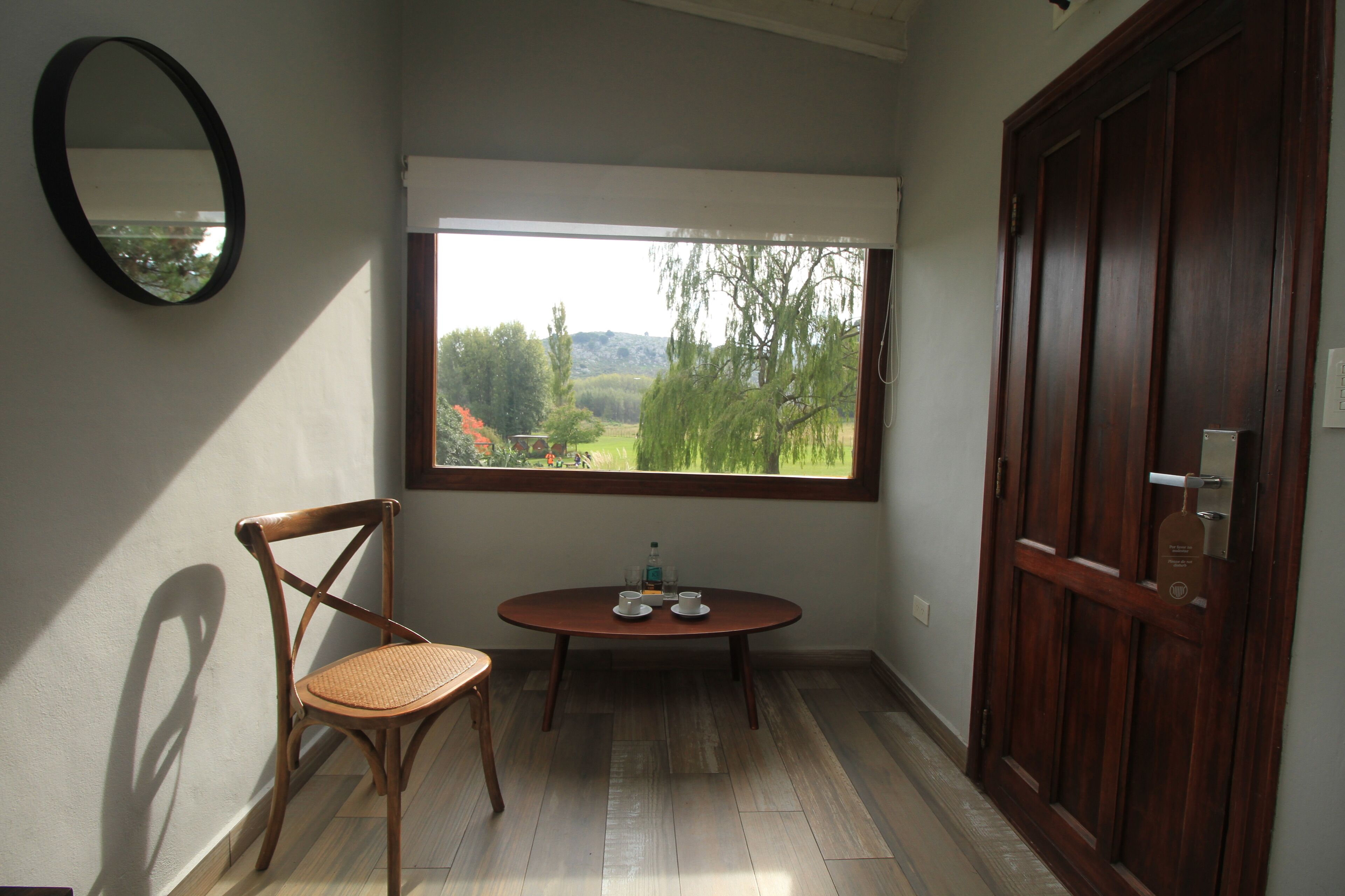 Una habitación con una silla de madera, una mesita con dos tazas, una ventana que ofrece vistas a árboles y colinas, y una puerta de madera.