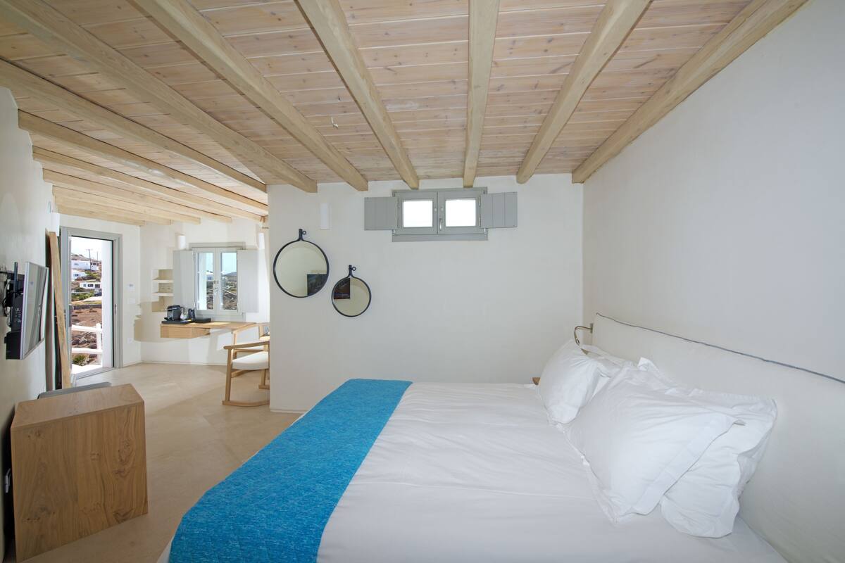 Quarto com teto de madeira, cama com roupa de cama branca, manta azul, escrivaninha com cadeira e dois espelhos fixados na parede.