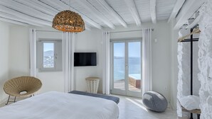 A bedroom with a bed, a wicker chair, a television, and a view of the sea through a sliding door.