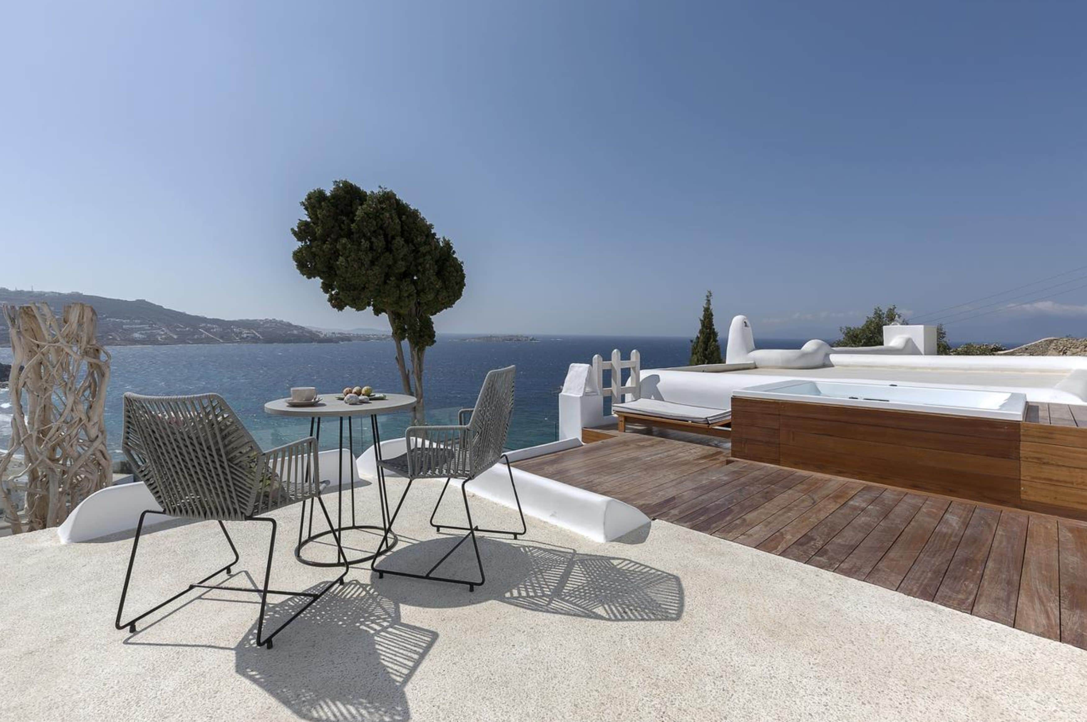 Un área de estar al aire libre con luz solar, una mesa, sillas, una terraza de madera y vista al mar.