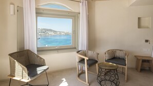 A room with a large window offering a view of a coastal town, two chairs, a small round table, and a wooden ceiling.