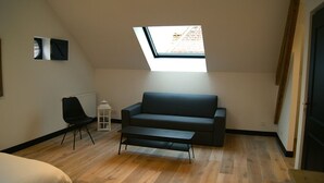 A loft bedroom with a sloped ceiling, a skylight, a dark sofa, a small table, and a chair. - La Grange de Salomé (Salomé)