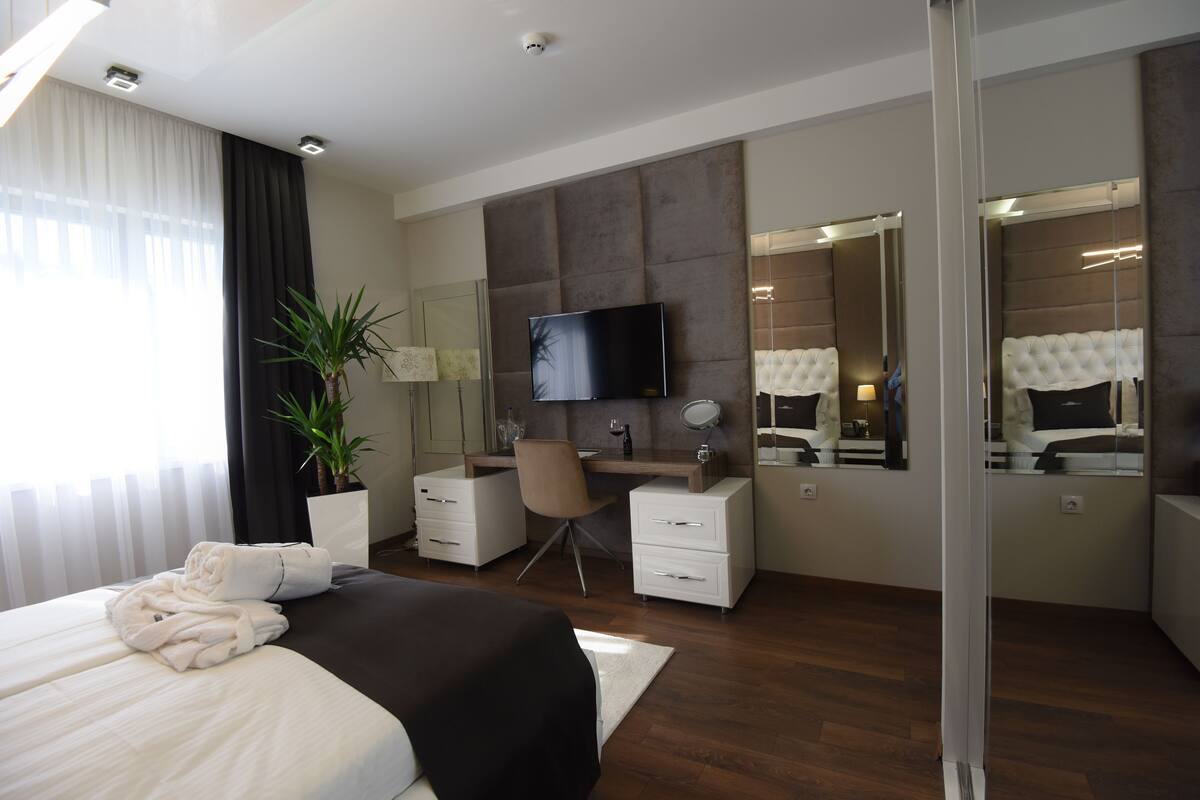A modern hotel room with a large bed, a desk, a chair, a mirror, and a flat-screen TV.