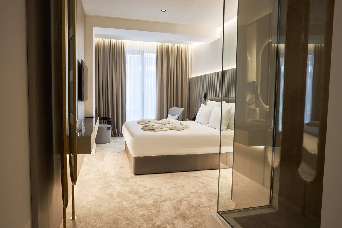 A modern hotel room with a large bed, a glass partition, and a view of the cityscape.