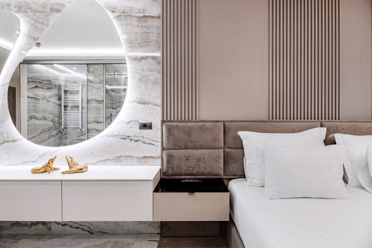 A modern bedroom with a marble vanity, a large mirror, a bed with white linens, and a built-in storage drawer.
