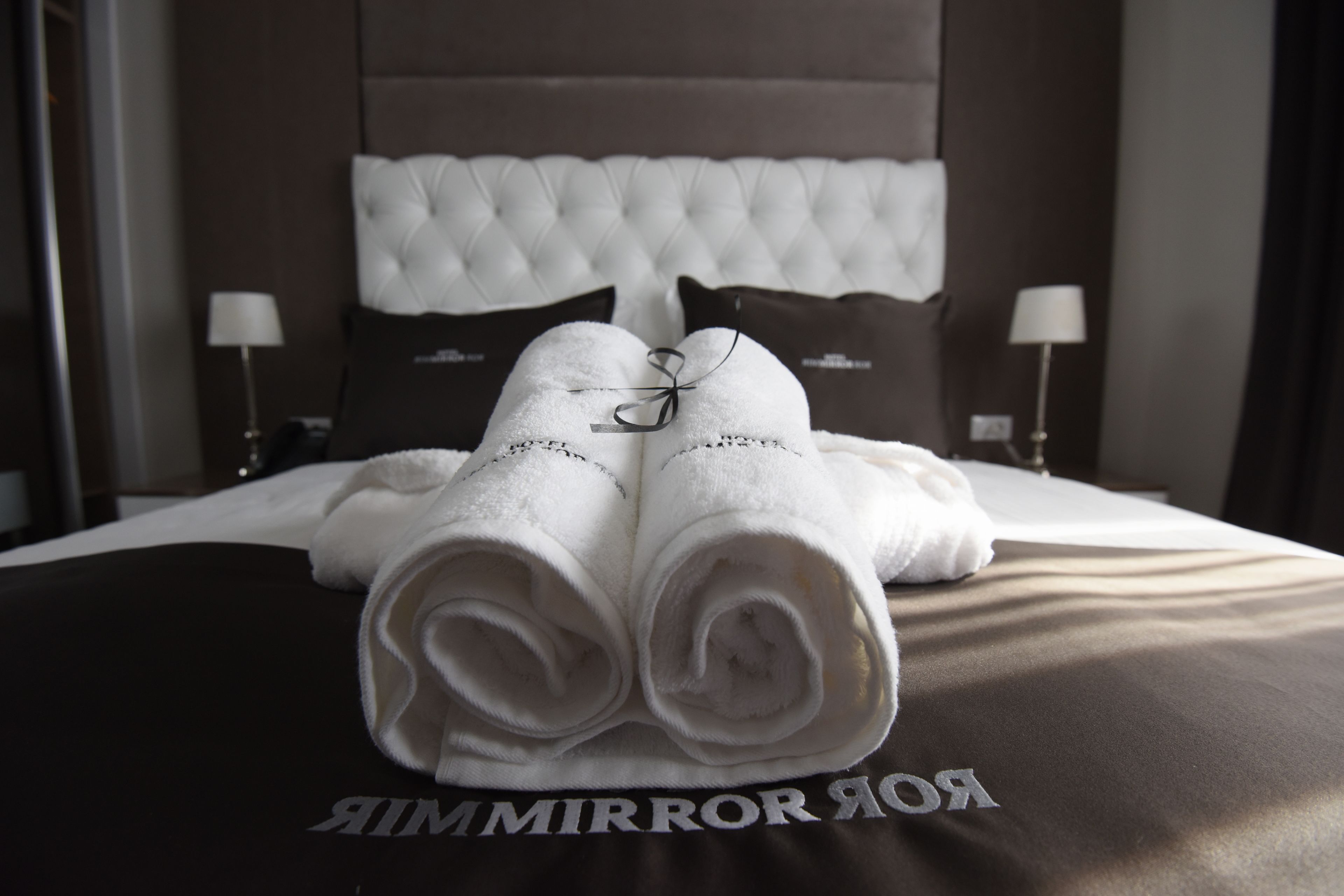 A neatly made bed with a tufted headboard, two pillows, and a pair of rolled towels on a dark bedspread with "RIMIRROR" text.