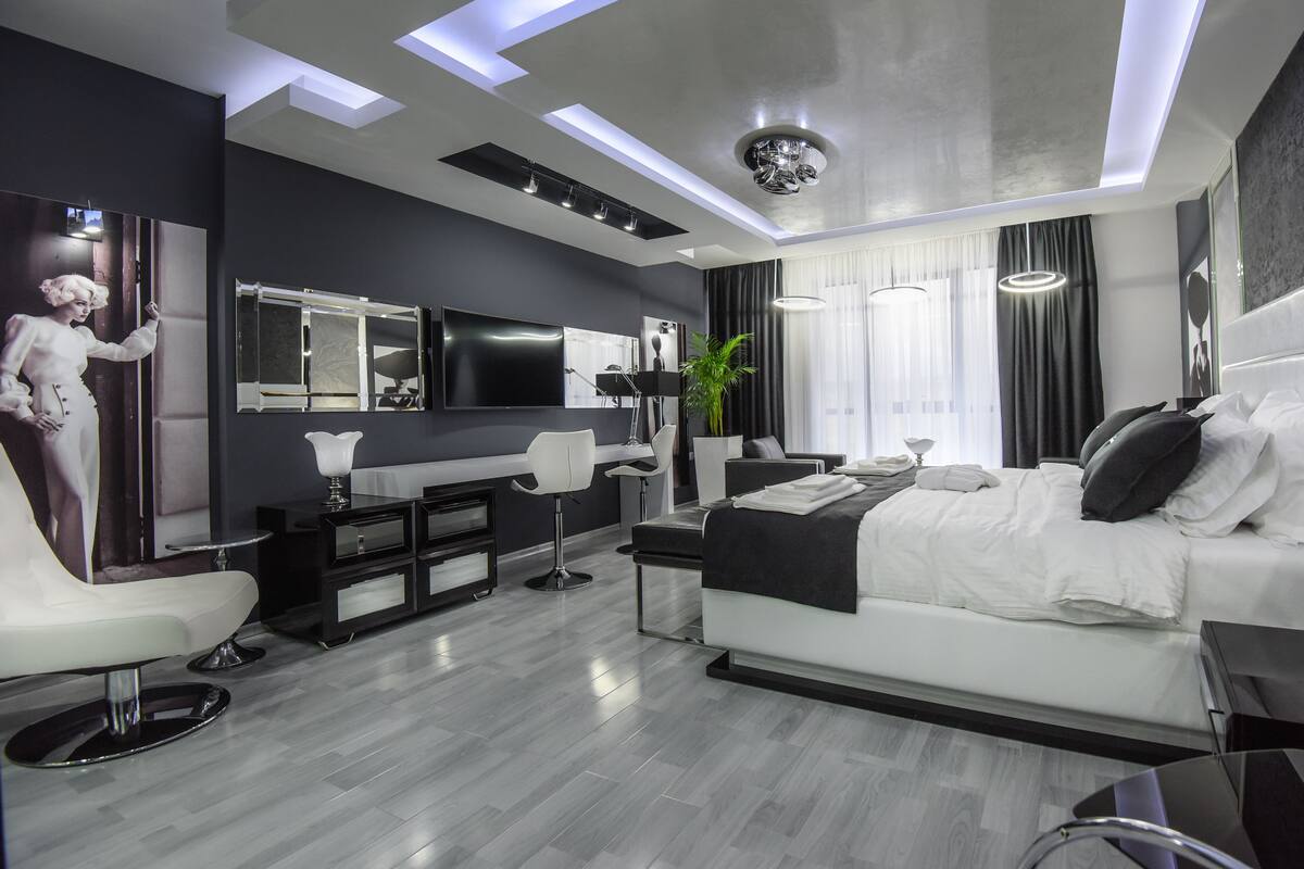 A modern hotel room with a large bed, a desk, a vanity with a mirror, a flat-screen TV, and a large window with curtains.