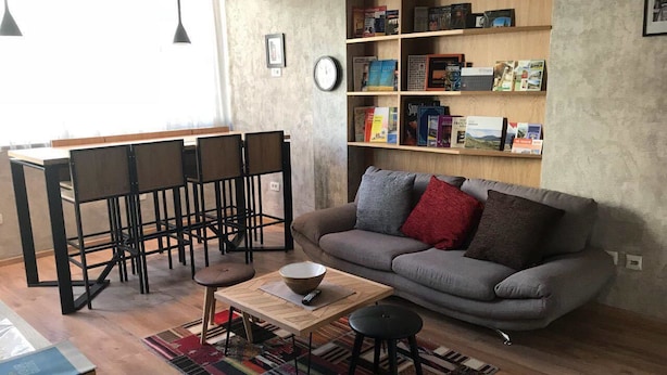 A modern living room with a grey sofa, a coffee table, a bookshelf, and a dining area with chairs and a table. - Hostel Boulevard Prishtina (Pristina)