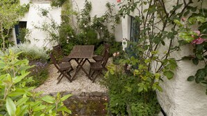 Zona d'estar a l'exterior amb un ambient rural, amb una taula i cadires de fusta, envoltada de vegetació abundant i plantes florides.