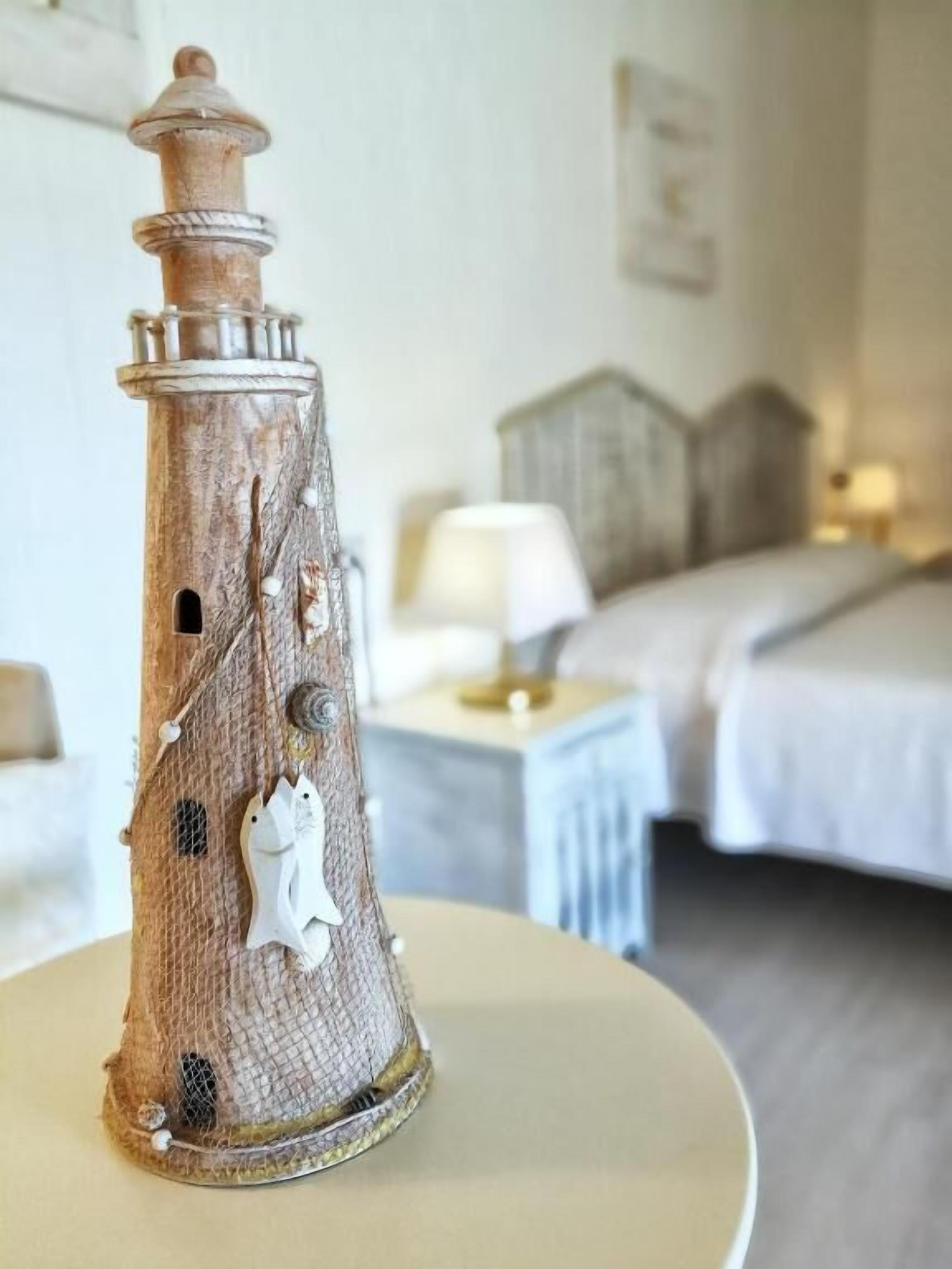 Un modelo rústico de faro sobre una mesa en una habitación de hotel, con una cama y una mesita de noche al fondo.