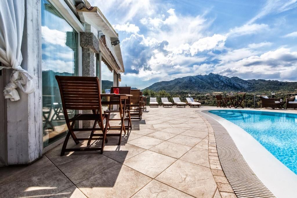 Zona de piscina con sillones reclinables y una mesa, con vistas a una cadena montañosa.