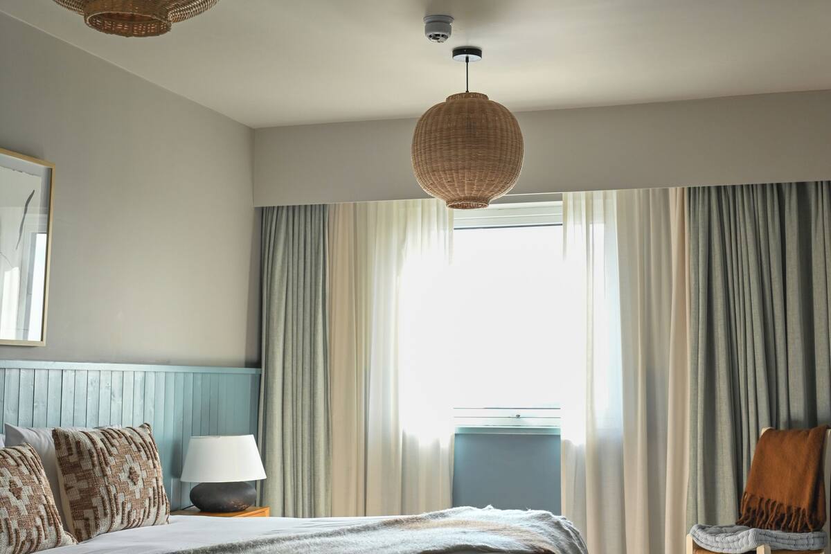 Quarto com cama grande, duas luminárias de vime suspensas, uma cadeira, luminária em mesa de cabeceira e janela com cortinas.